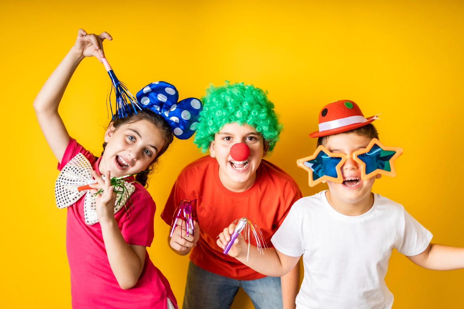 Three children with party props including wig, party hats and star glasses on yellow