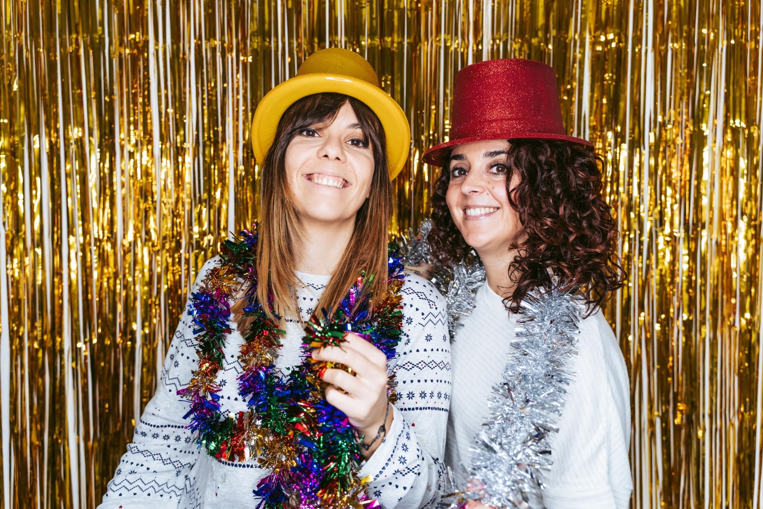Two women in fedora hats with tinsel boas posing at gold metallic curtain backdrop